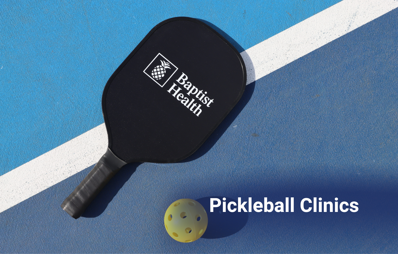Pickleball paddle with the Baptist Health logo and a yellow pickleball resting on a blue court surface.