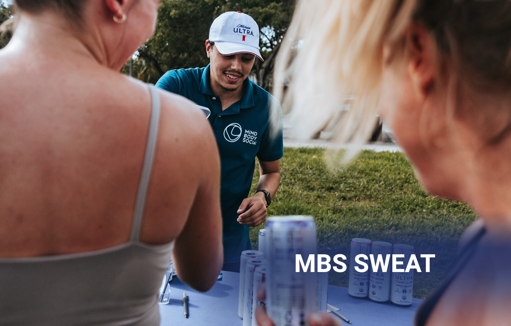 Event staff member handing out beverages at an outdoor MBS Sweat wellness event with participants gathered around the table.
