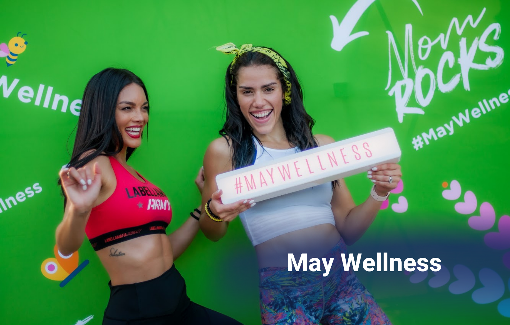 Two women posing enthusiastically in front of a green May Wellness event backdrop while holding a hashtag sign.
